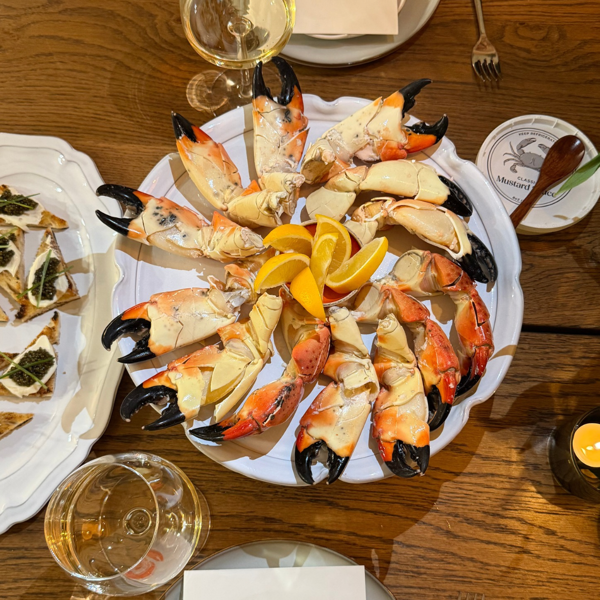 Plate of stone crab claws organized in a circle, with a pile of lemon wedges placed in the middle. There is a plate of chips with creme fraiche and caviar on top on the side with two glasses of wine and a pot of mustard sauce on the table.