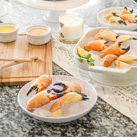 A spread of stone crab claws on ice served in white bowls, accompanied by dipping sauces, wooden crab mallets, and garnished with lemon and herbs on a lace-covered table