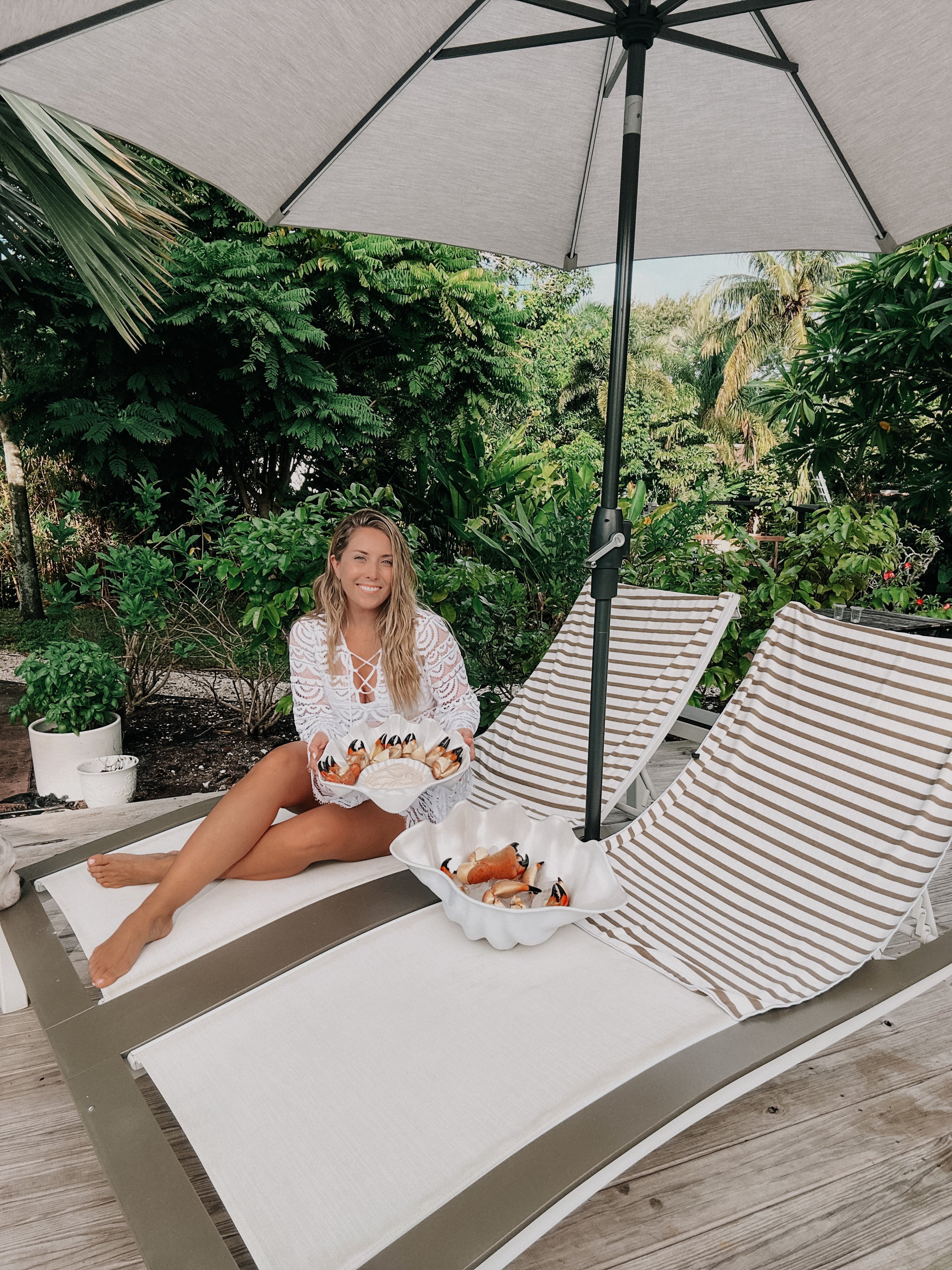 Woman in white cover-up holding a bowl of stone crab claws on a lounge chair under a patio umbrella, surrounded by lush tropical greenery
