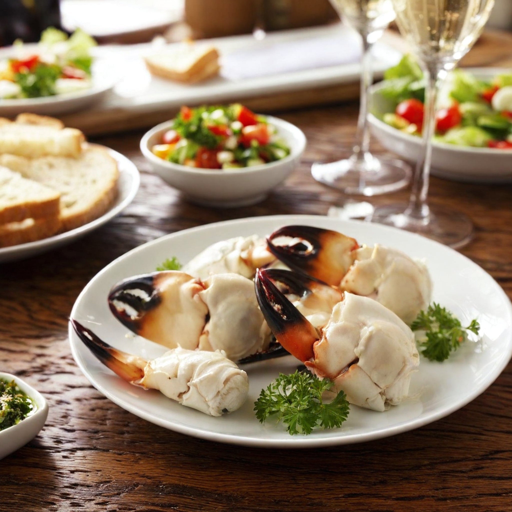 Stone crab claws arranged on a white plate with fresh parsley on a wood table with bread, salad, and glass flutes