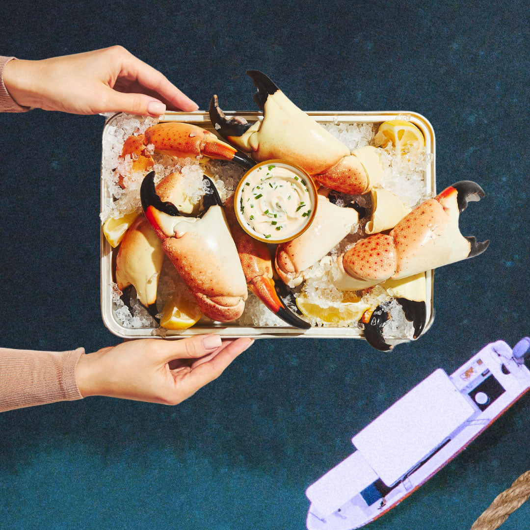 Hands holding a tray of stone crab claws on ice with lemon wedges and dipping sauce, over a dark blue background with part of a boat visible